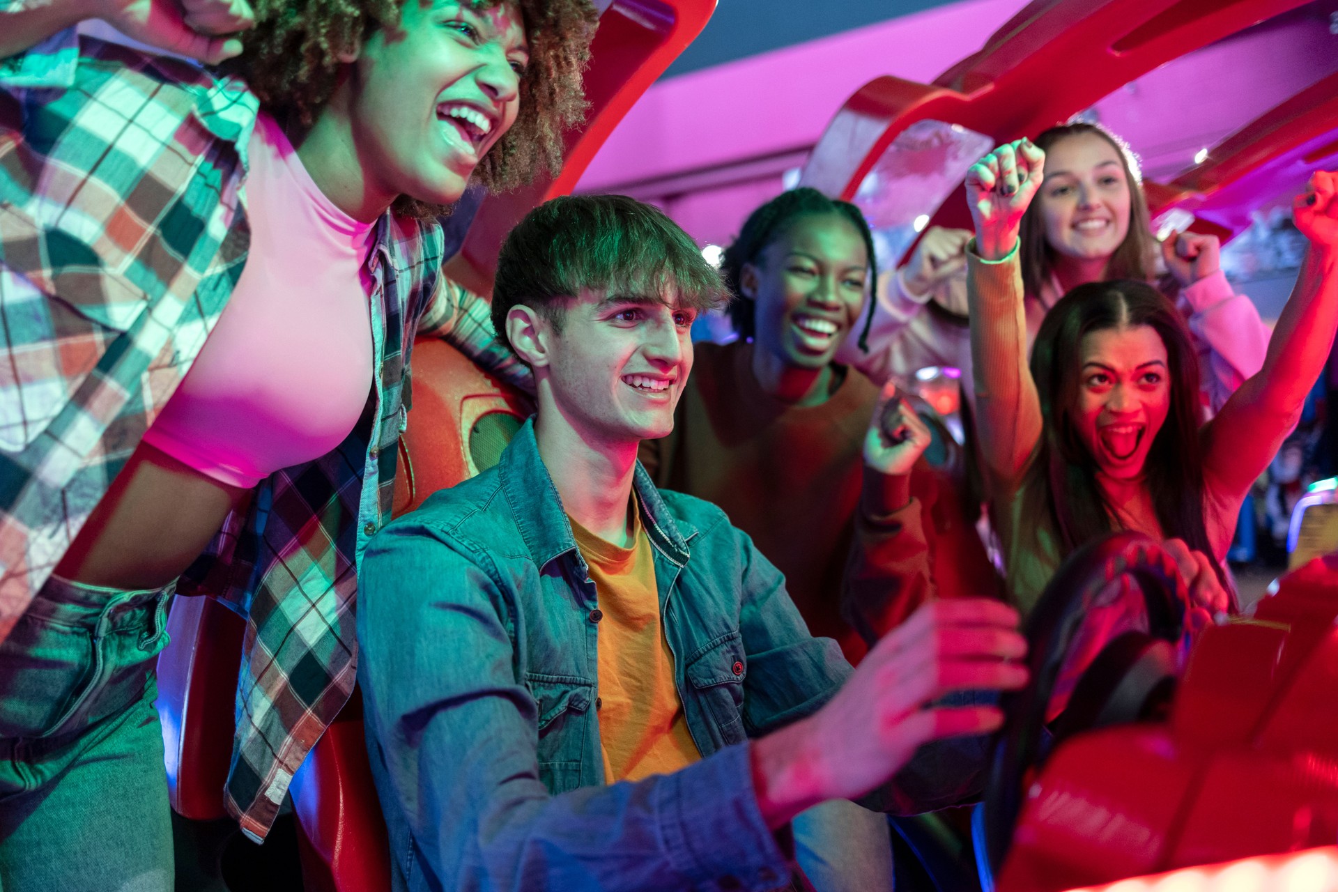 group of multiracial gamer friends playing racing arcade machines while celebrating excitedly